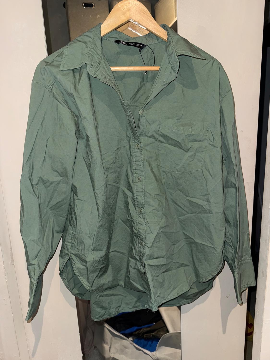 Wrinkled green shirt hanging on a wooden hanger, with a white wardrobe background and partially visible objects.
