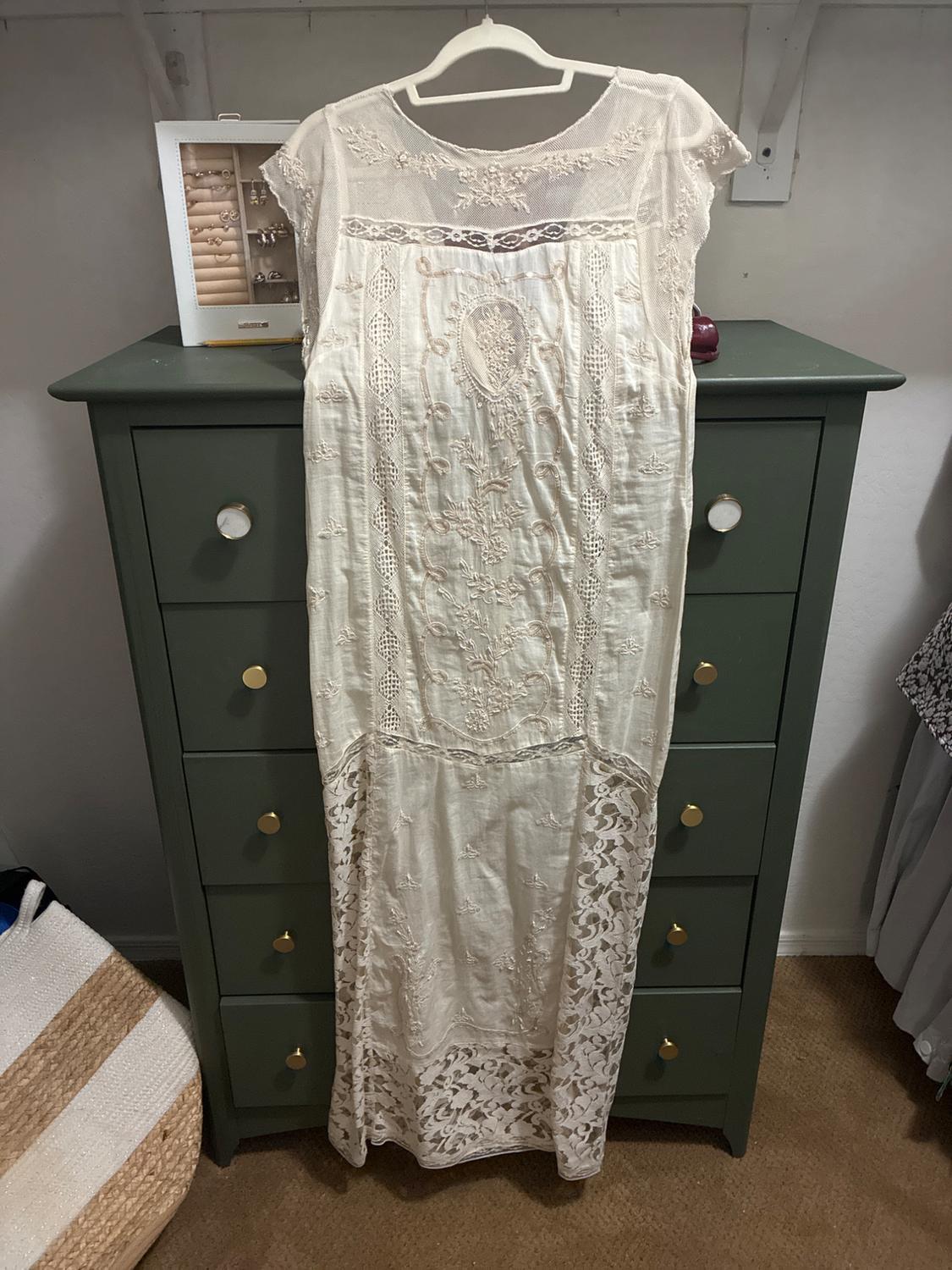 Cream-colored long dress with embroidery and lace details, hanging on a hanger in front of a dark green dresser.