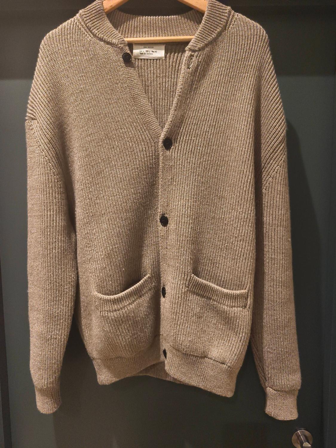 Beige knitted cardigan with black buttons and front pockets, hanging on a wooden hanger against a dark background.
