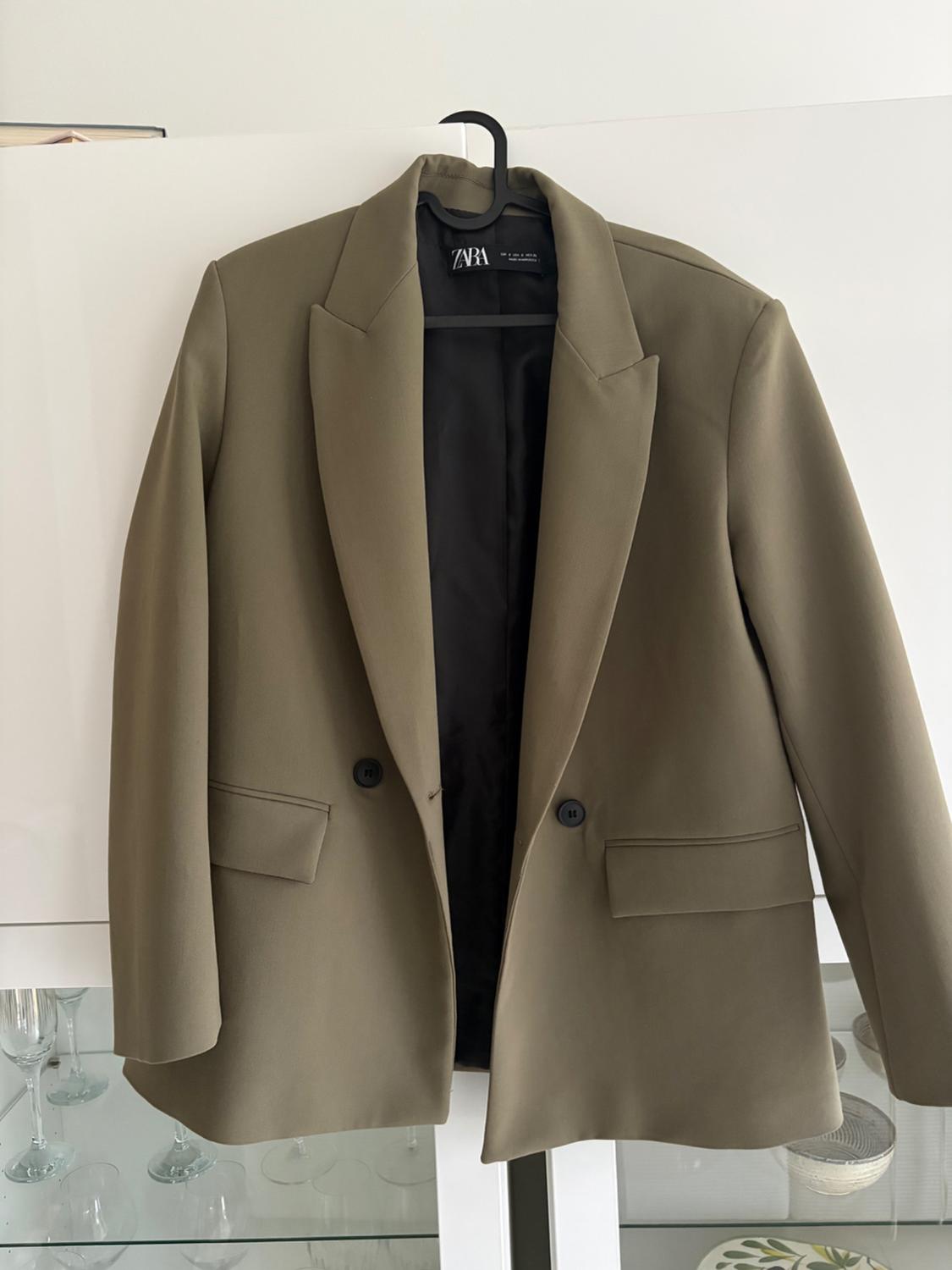 Olive green jacket hanging on a black hanger, with two buttons and front pockets. Visible label: "ZARA". White background with crystal glasses.