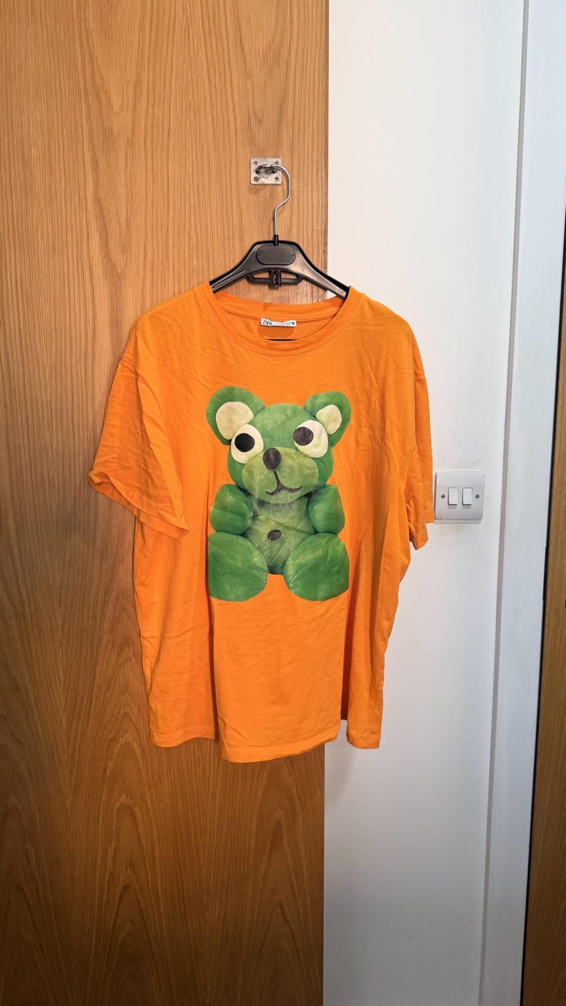 ZARA orange T-shirt with a green bear print, hanging on a hanger over a wooden door in a relaxed setting.