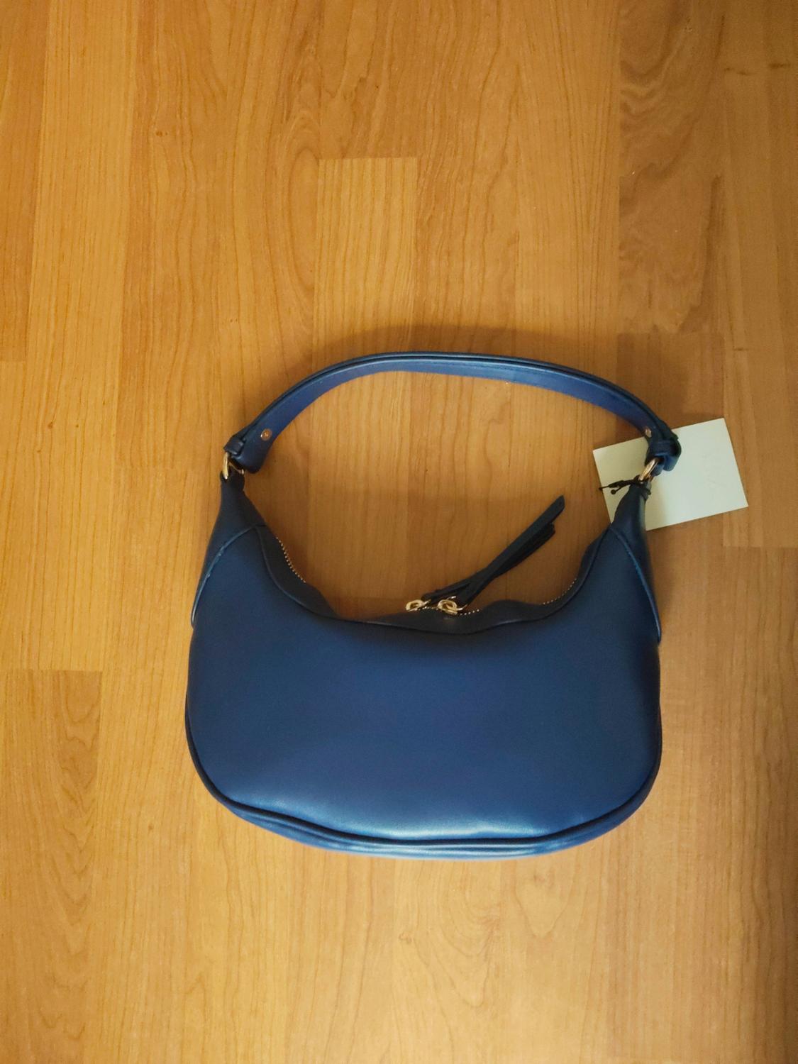 Dark blue shoulder bag with gold detailing and a label, resting on a wooden floor.