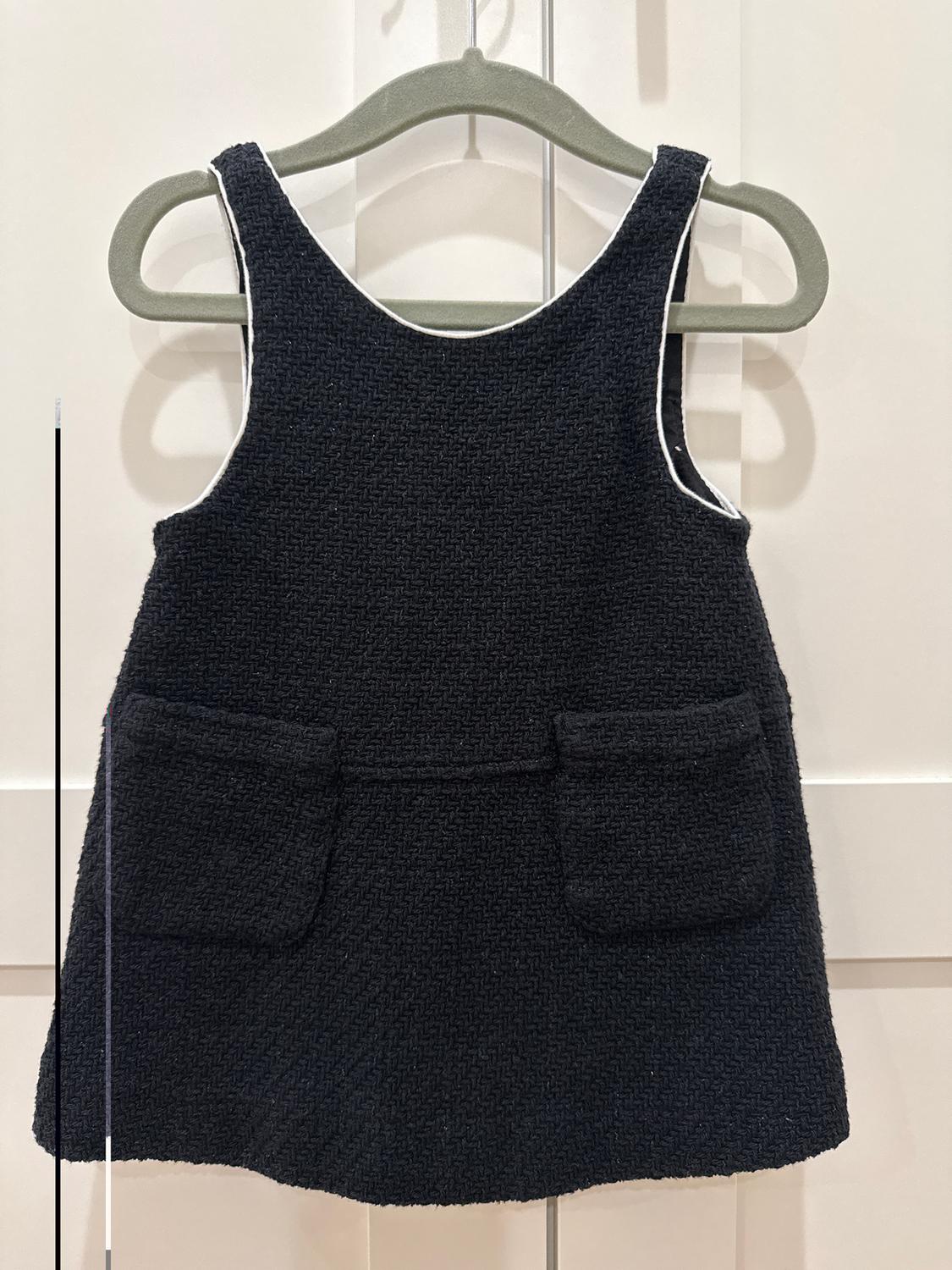 Black children's dress with a woven texture and front pockets, hanging on a grey hanger against a white background.