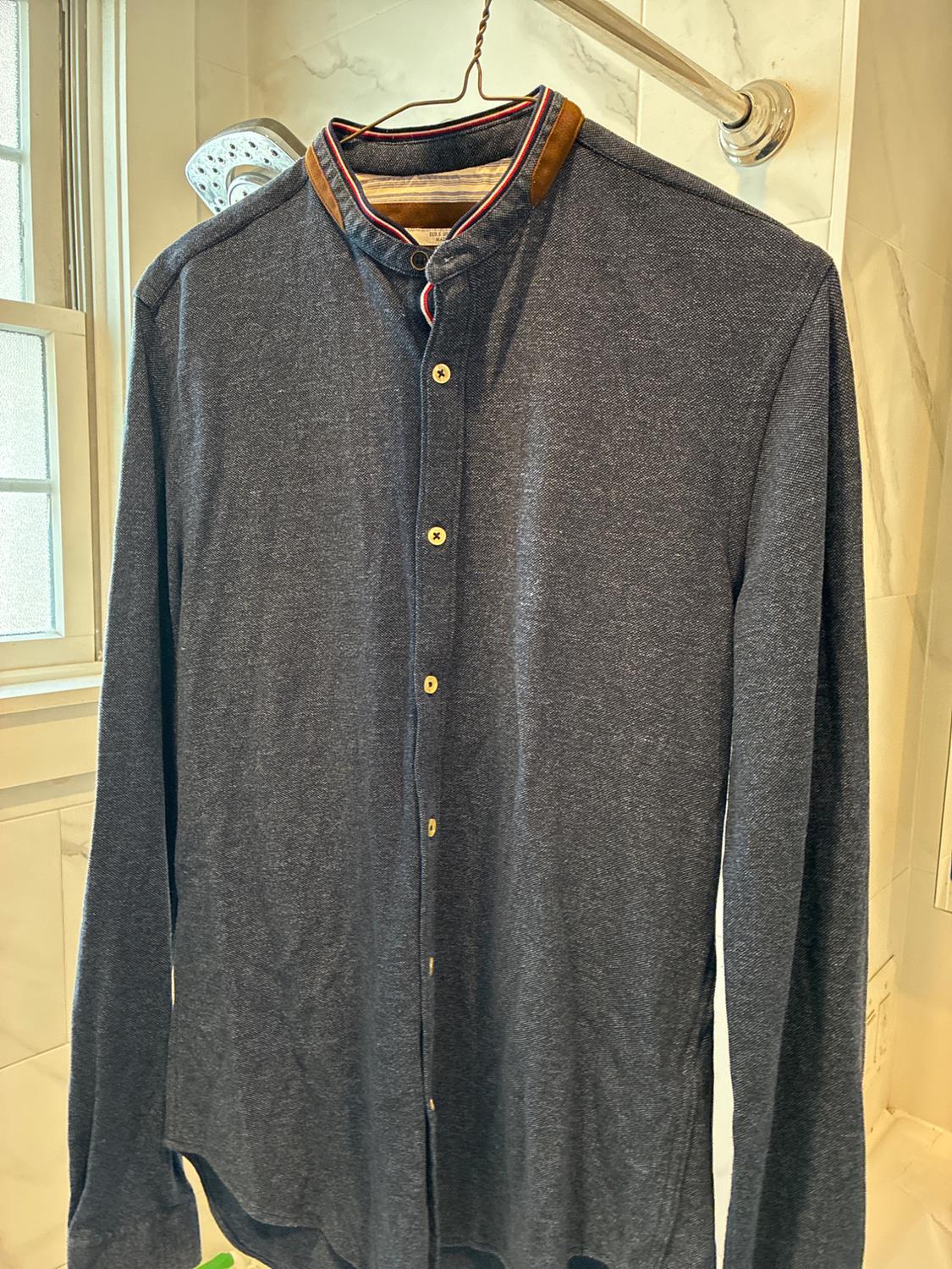 Long-sleeve blue shirt with a mandarin collar and buttons, hanging on a wire hanger in a bathroom with white tiles and a shower.