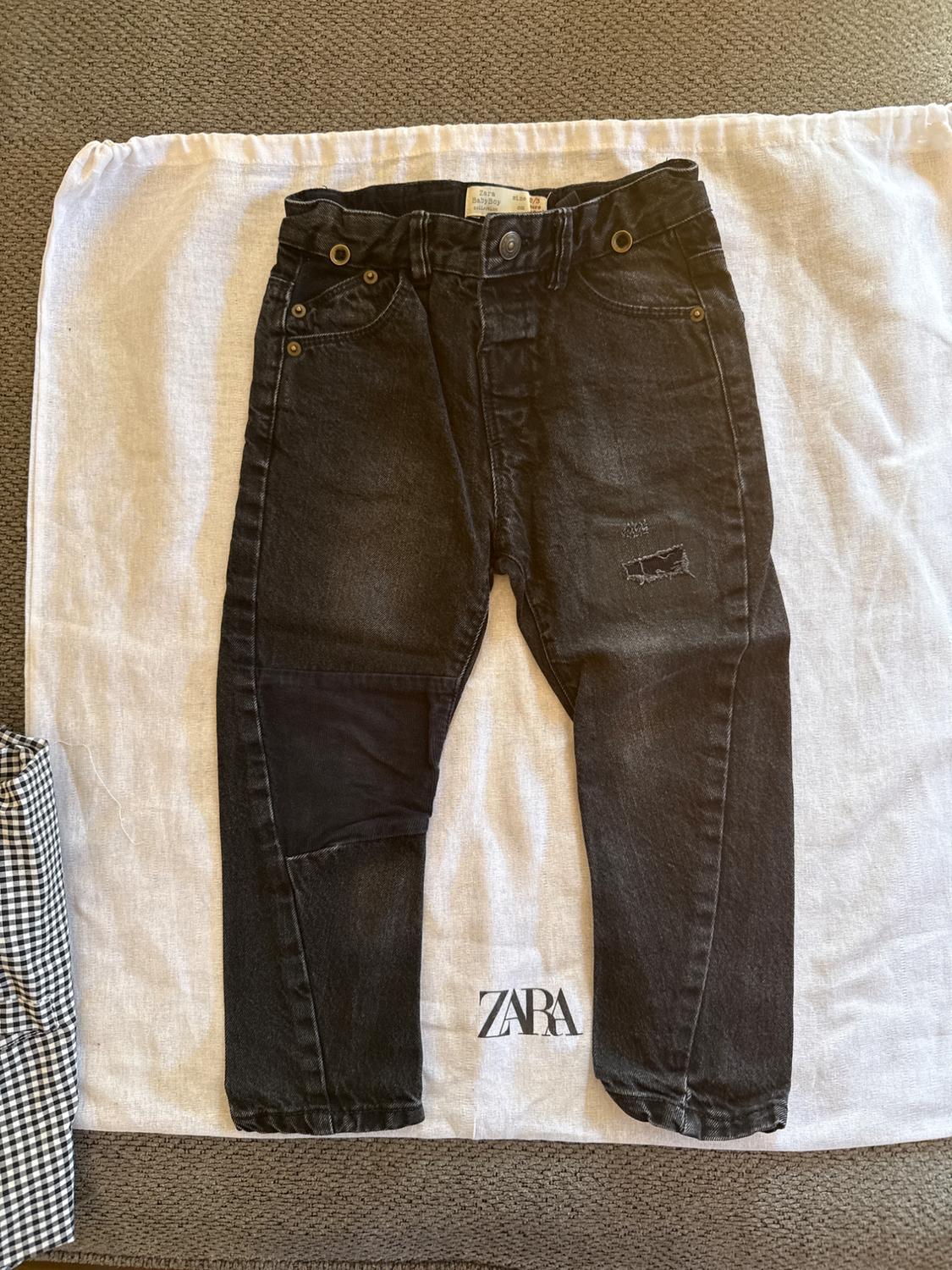 Black denim trousers with patches, on white fabric with "ZARA" logo. Grey fabric background and checked fabric detail.