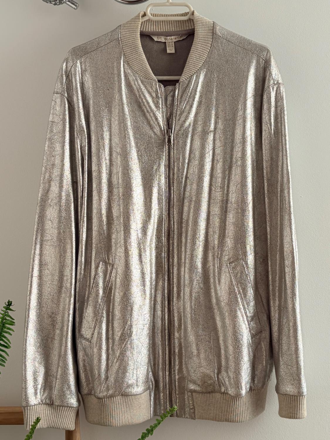 Silver metallic jacket hanging on a beige hanger, with a zip and pockets, on a white background. Visible label: "trf outerwear".