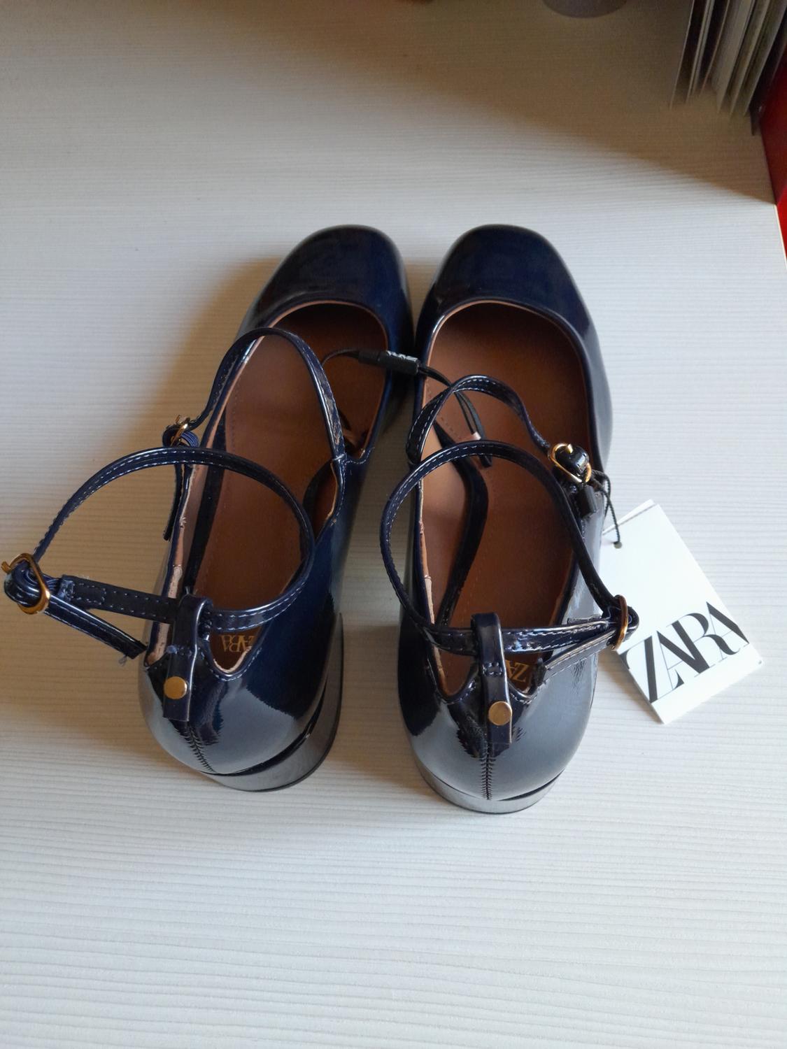 Navy blue patent leather high-heeled shoes with an ankle strap on a light-coloured surface. White label with the text ZARA.