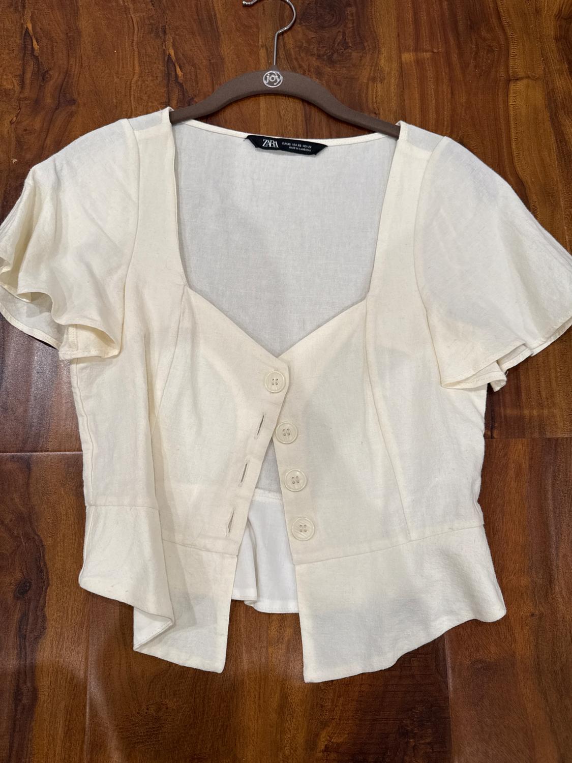 Short-sleeved white blouse with large buttons, hanging on a hanger against a brown wooden background. Visible label: "Zara".