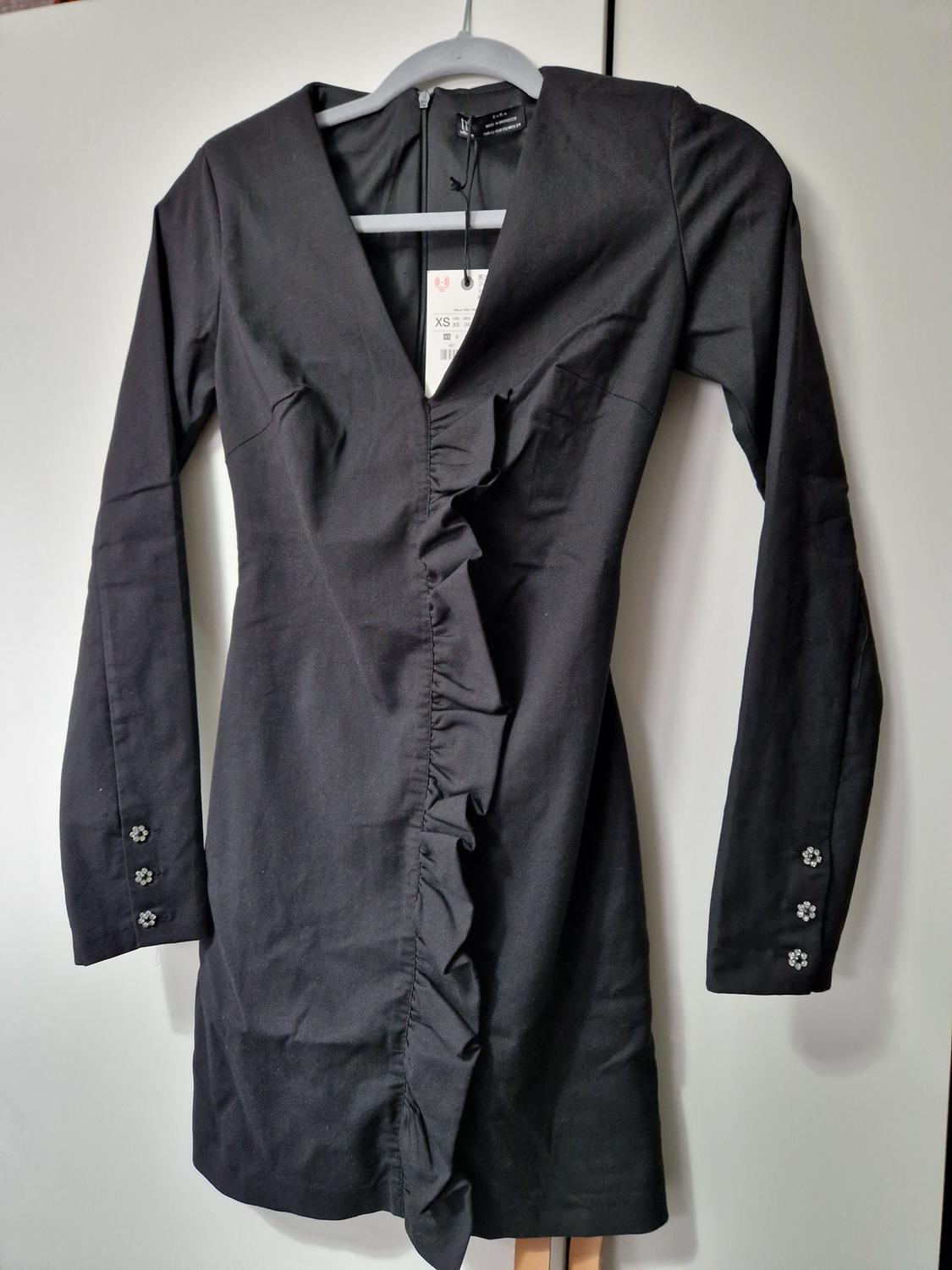Long-sleeved black dress, hanging. V-neck, front frills and buttons on the cuffs. Label with size XS.