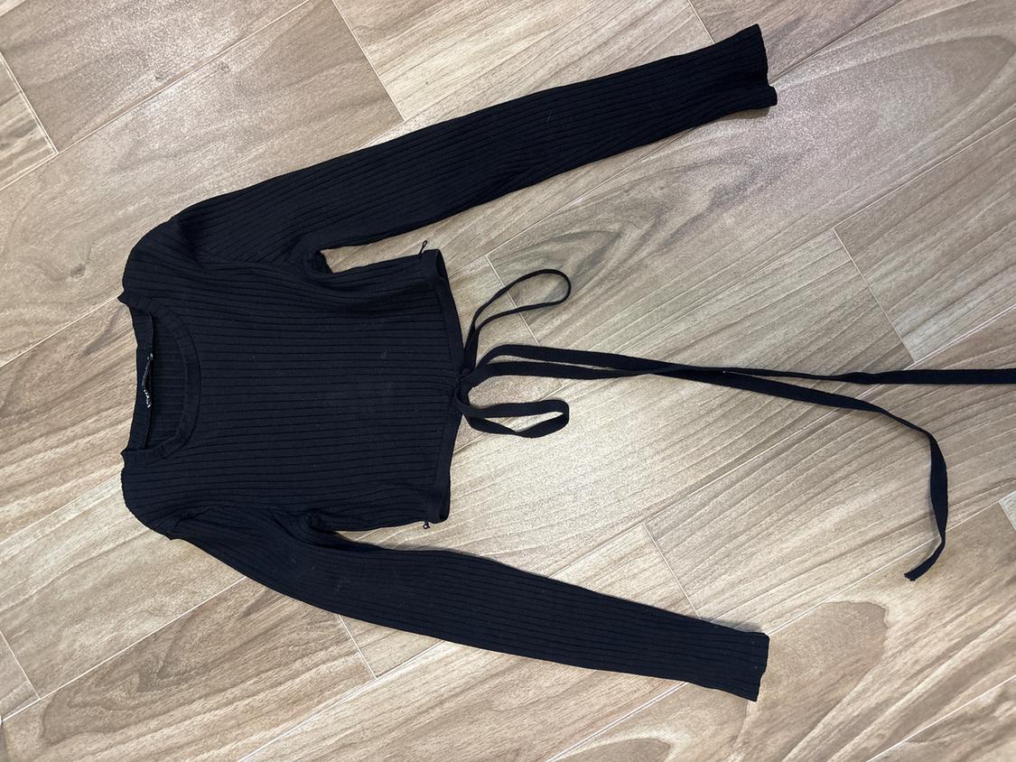 Black long-sleeved ribbed top with long waist ties, on a light wood floor.