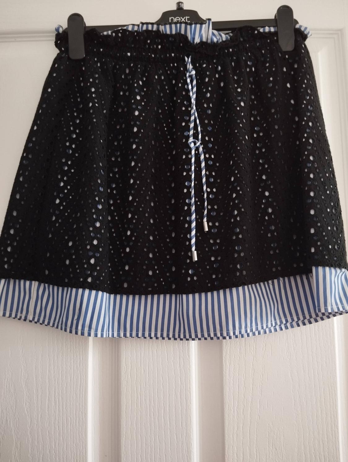 Black perforated skirt with a blue and white striped hem hanging on a hanger on a white door. Text: next 16.