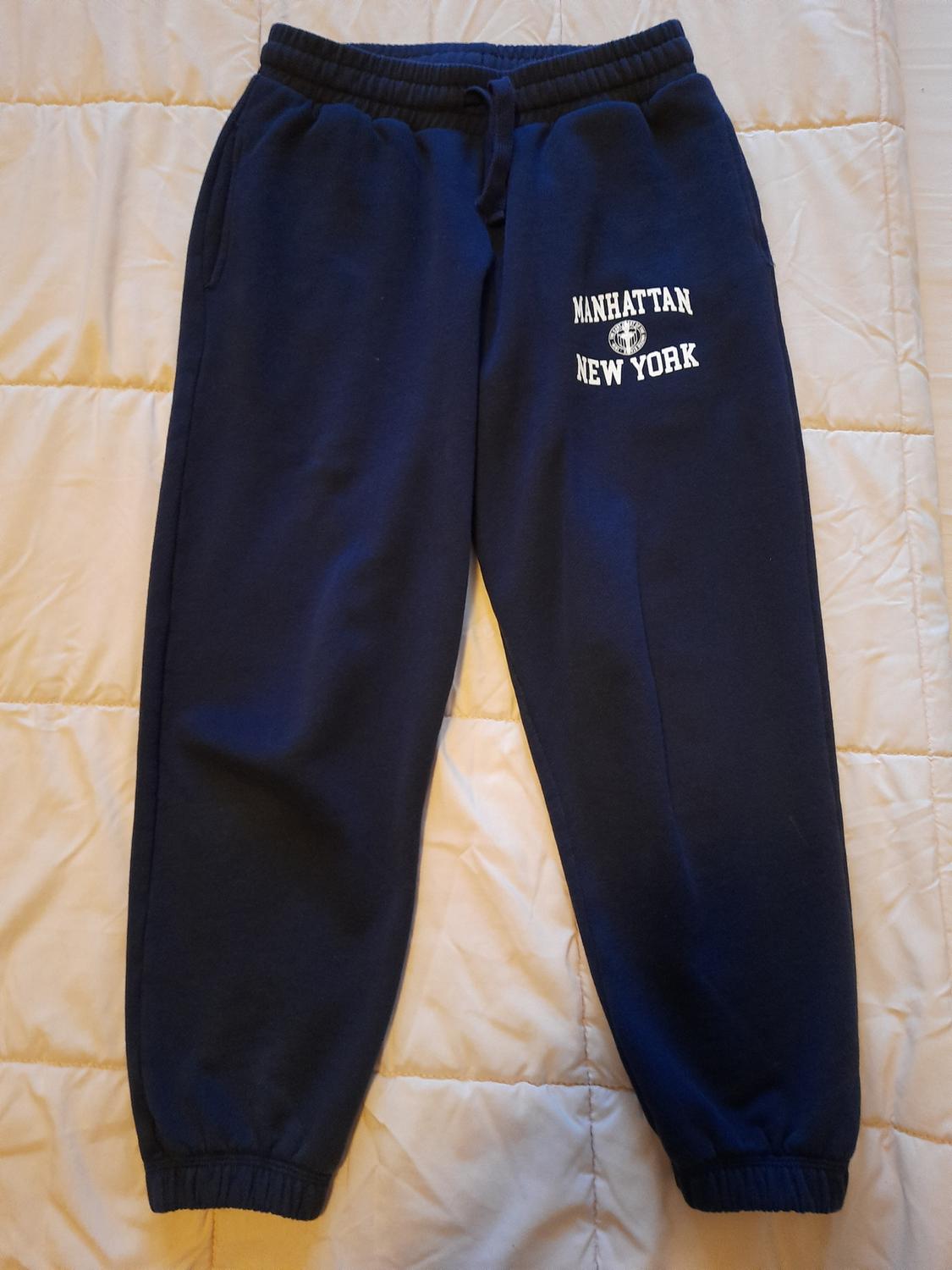 Navy tracksuit bottoms with "MANHATTAN NEW YORK" text in white, on a quilted beige bedspread.