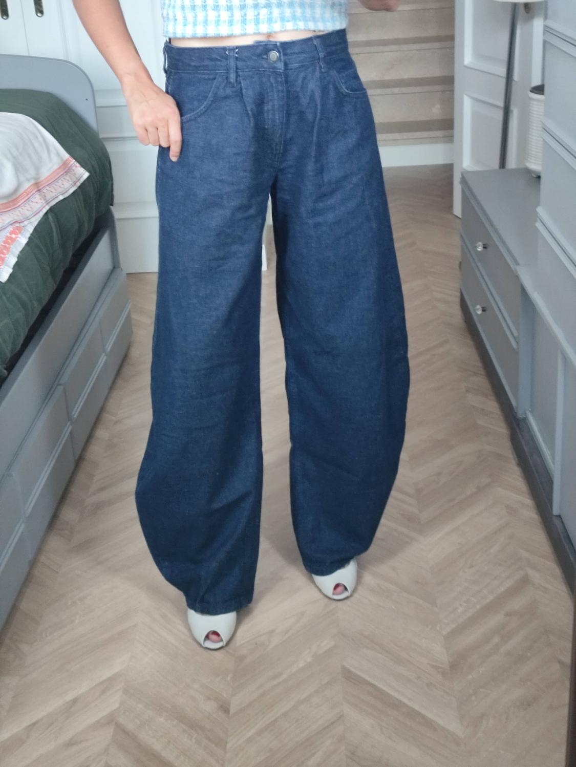 Person in a bedroom wearing dark blue baggy jeans, a light blue checkered crop top and white shoes. Wooden floor.