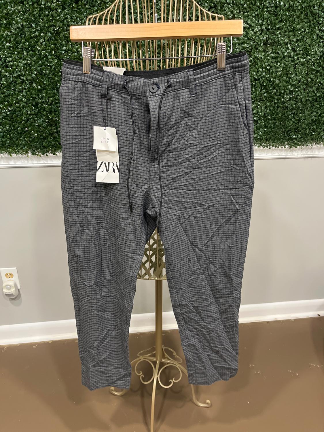 Gray pants with a plaid print, hanging on a golden hanger against a background of green leaves. Visible tags: "JOIN LIFE", "ZARA".