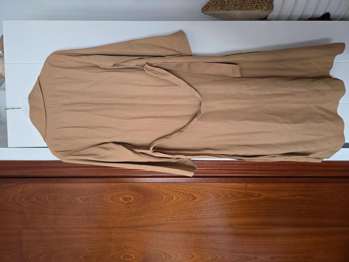 Long beige coat hanging on a wooden door. The coat has a belt of the same colour and is against a white background.