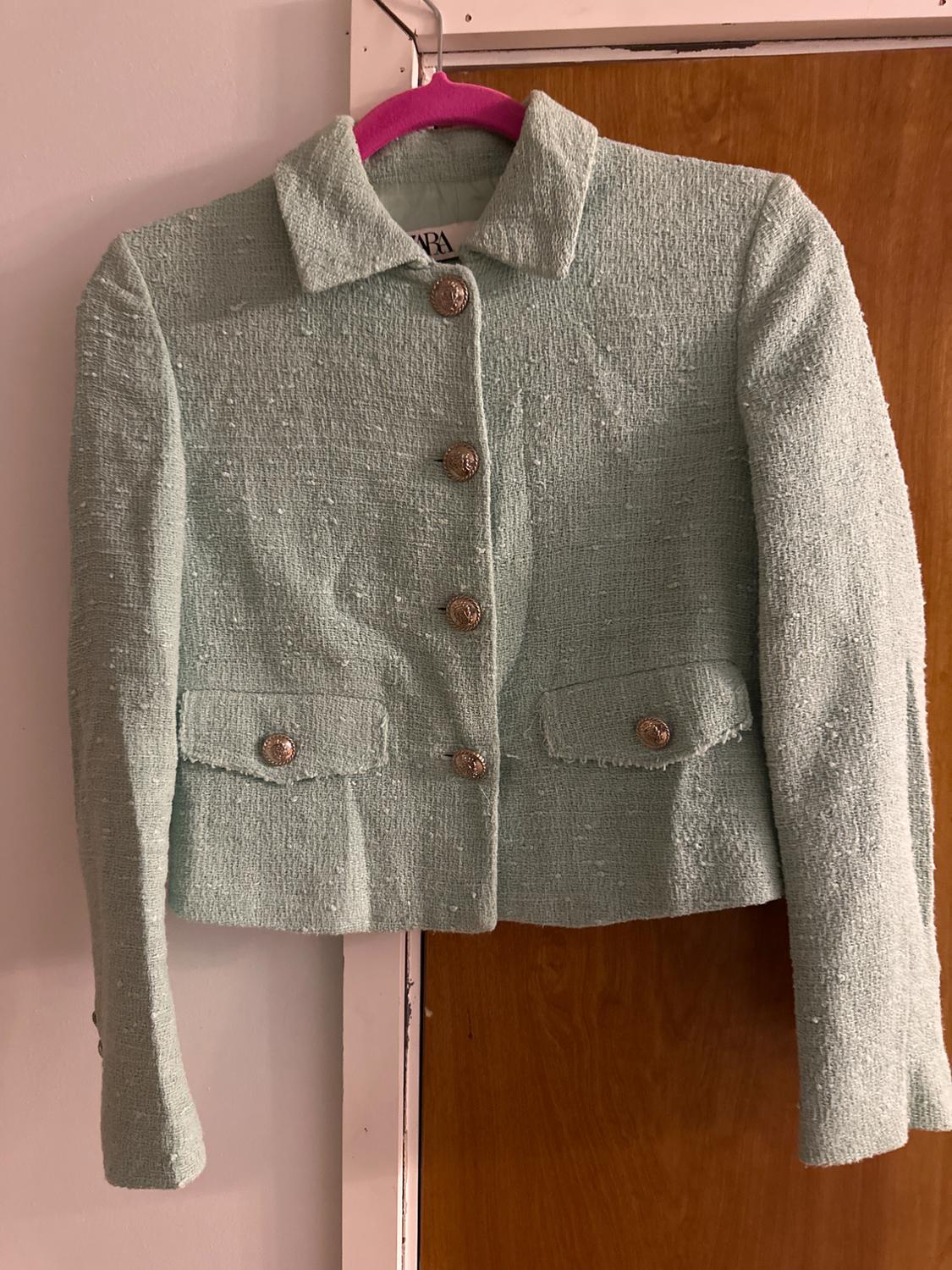 Light green textured jacket with metallic buttons and pockets, hanging on a pink hanger in a wooden doorway.