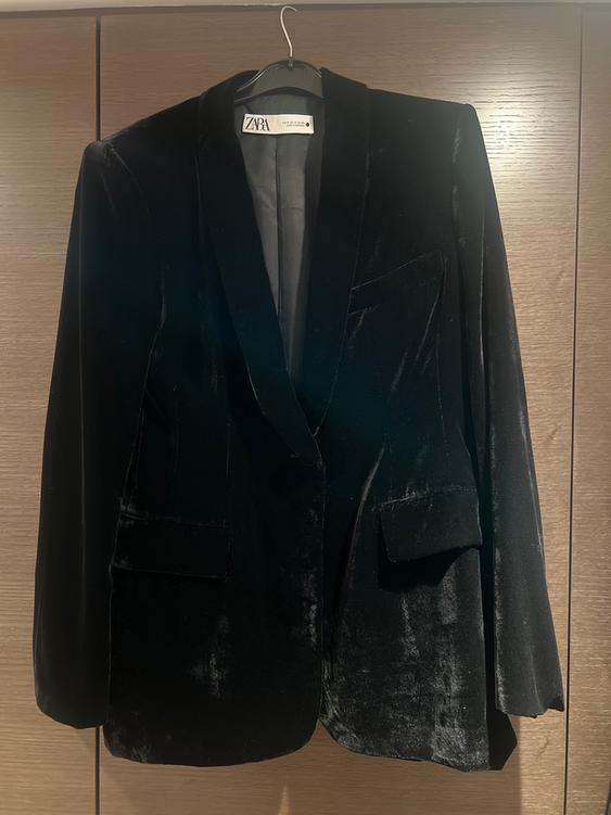 Black velvet jacket hanging on a hanger, with wide lapels and front pockets. Visible label: "ZARA". Light wood background.