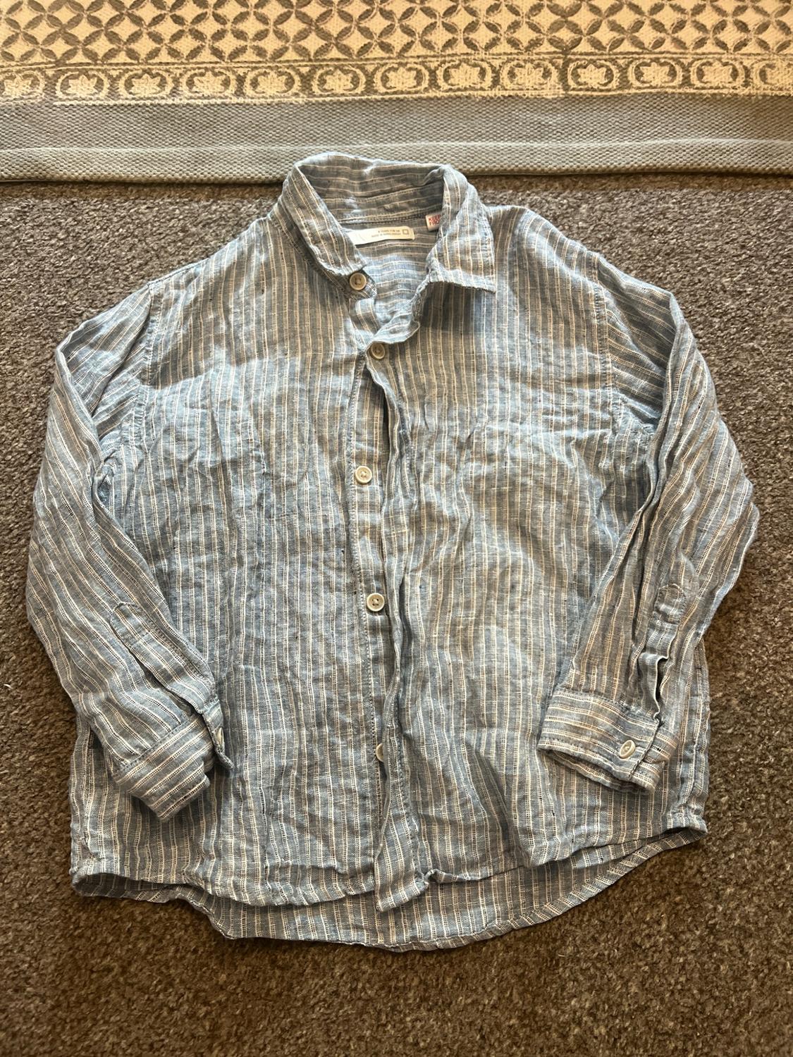 Blue and white striped long-sleeved shirt, buttoned up and laid out on a textured brown rug. Casual style.