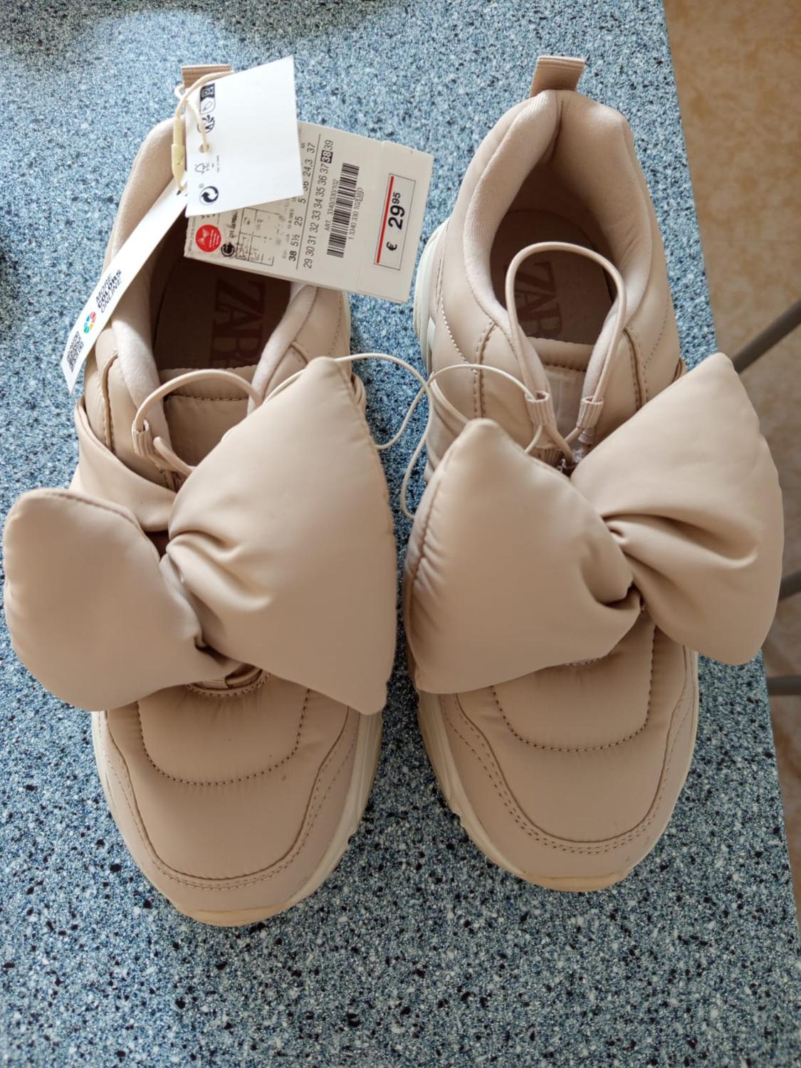 Beige slippers with a large bow on the top, on a blue speckled surface. Price tag of €29.95.