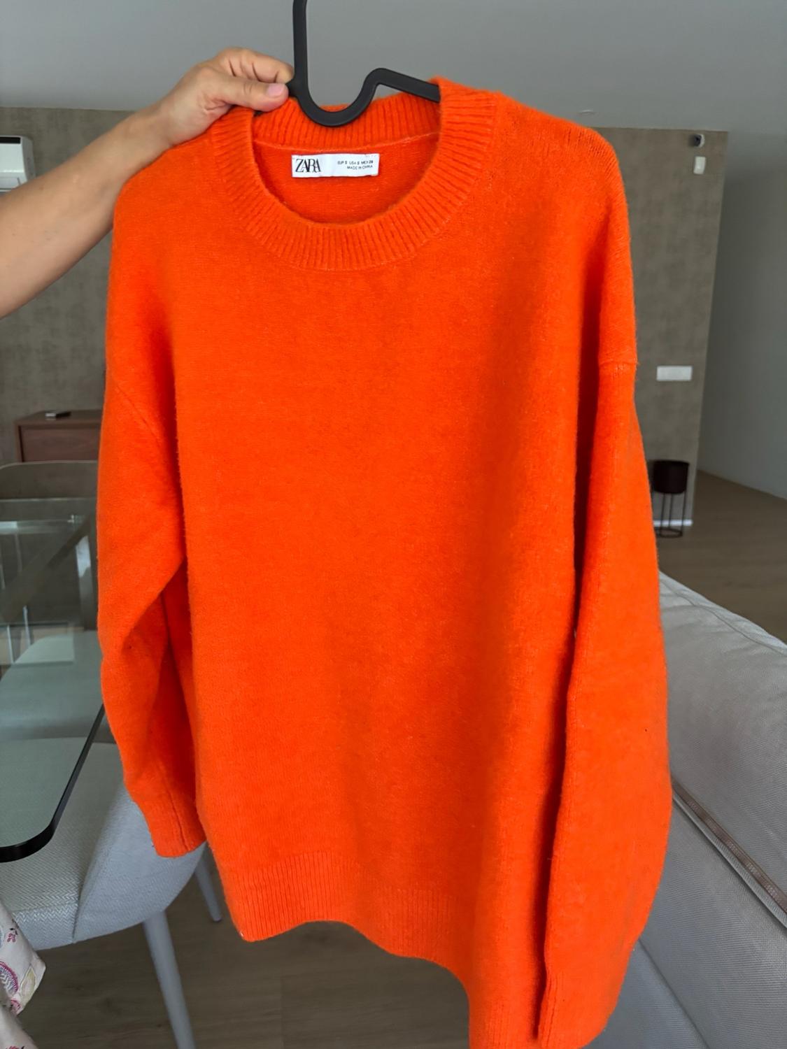 Hand holding an orange knit jumper on a black hanger. ZARA label visible. In the background, a living room with a glass table and sofa.