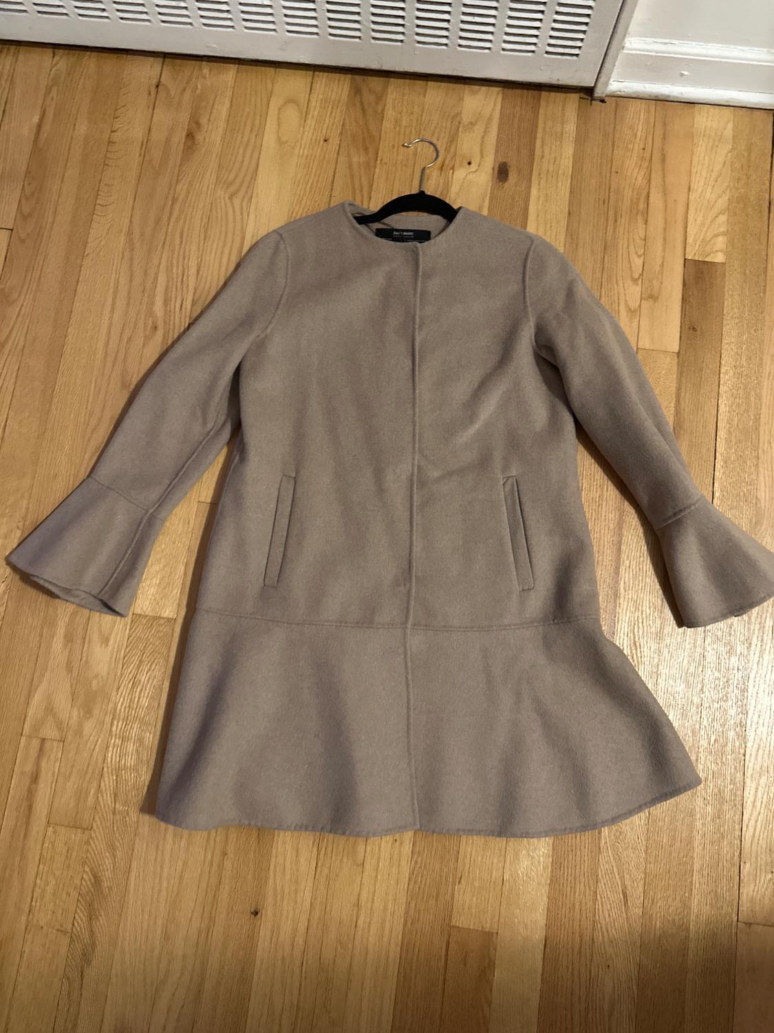 Beige wool coat with flared sleeves and a straight cut, hanging on a hanger over a light wood floor.