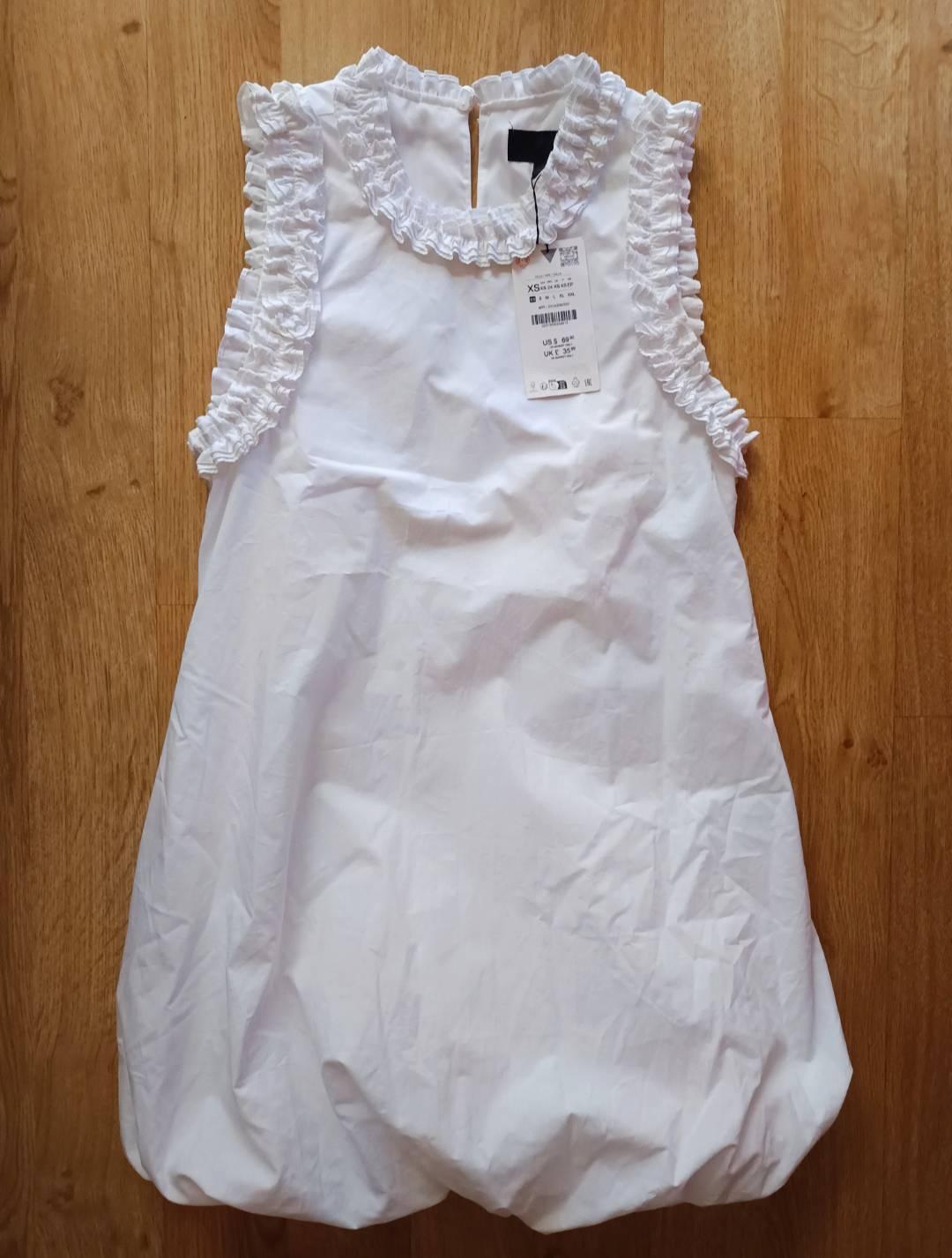 White sleeveless dress with frills at the collar and armholes, and a puffed hem, on a wooden floor. Label with text XS.