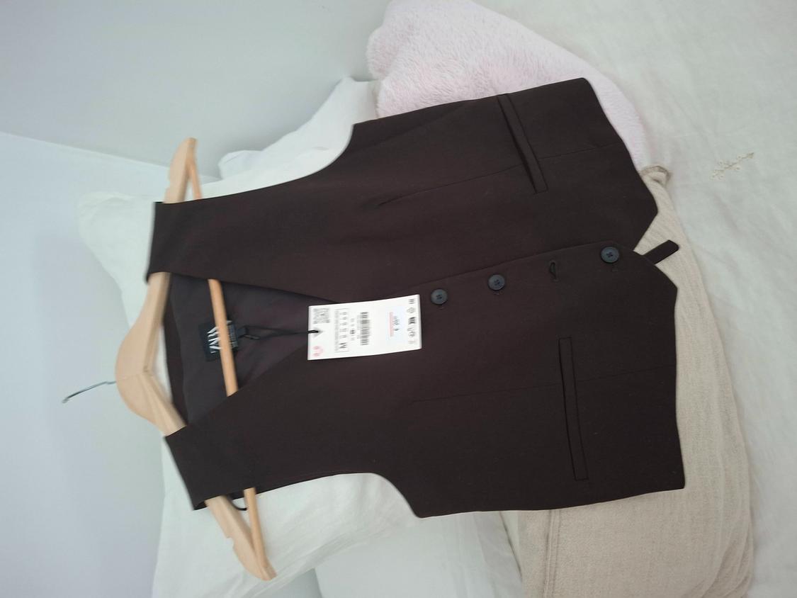Dark brown tailored waistcoat on a wooden hanger over a bed with white sheets. Label with ZARA text visible inside.