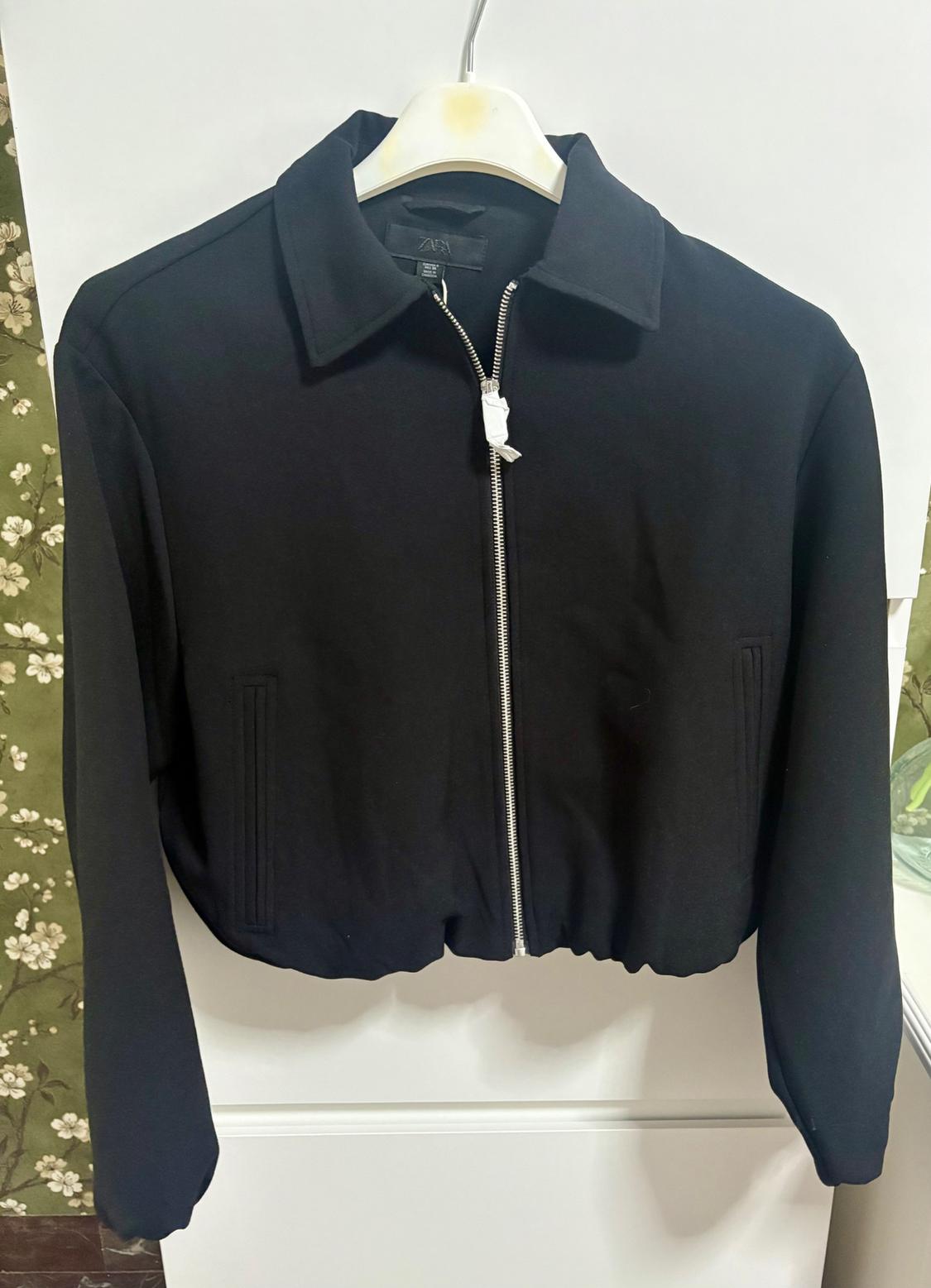 Black jacket with silver zip and classic collar, hanging on a white hanger. Background with green floral wallpaper.