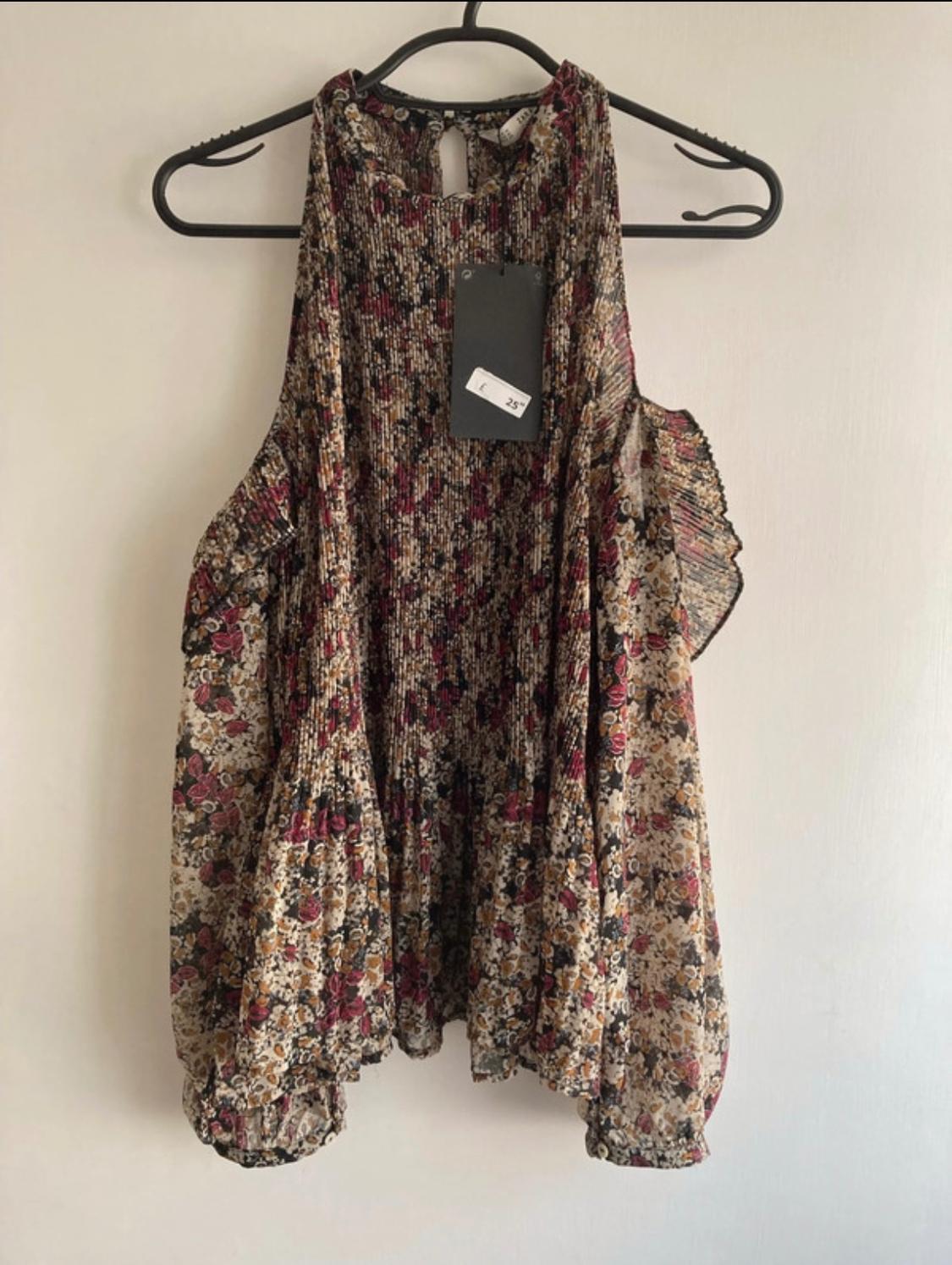 Front view of a sleeveless blouse with a floral print and pleats, hanging on a hanger.