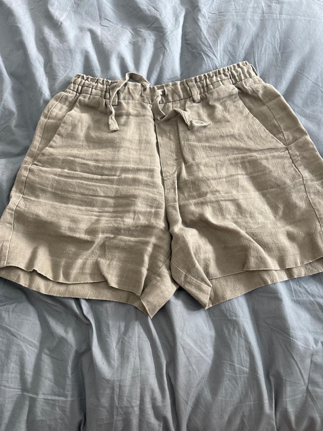 Beige shorts with an elasticated drawstring waist, on a crumpled grey fabric surface.