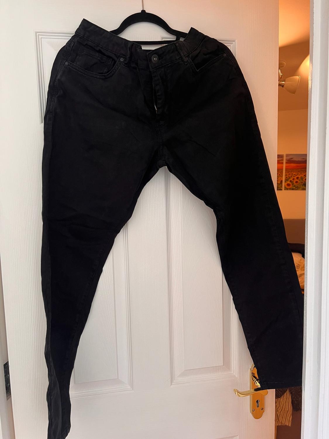 Black jeans hanging on a hanger on an ajar white door, with a bedroom in the background. Casual and relaxed style.