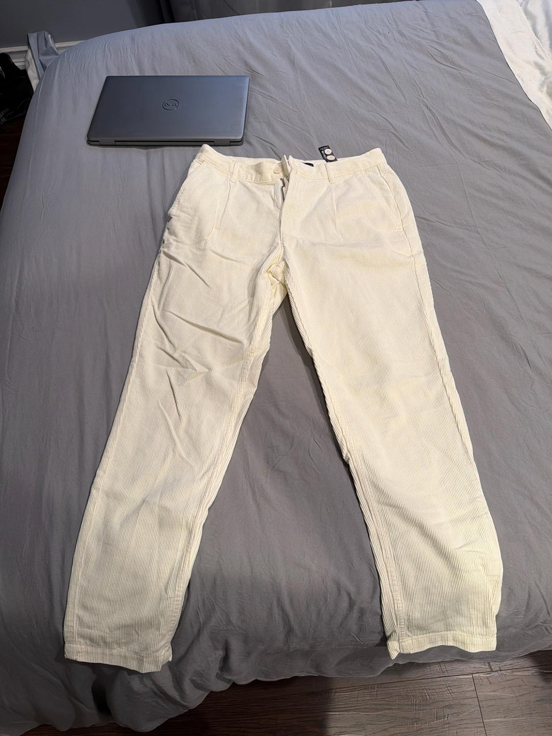 Cream-coloured corduroy trousers laid out on a bed with grey sheets next to a closed laptop. Casual and relaxed atmosphere.