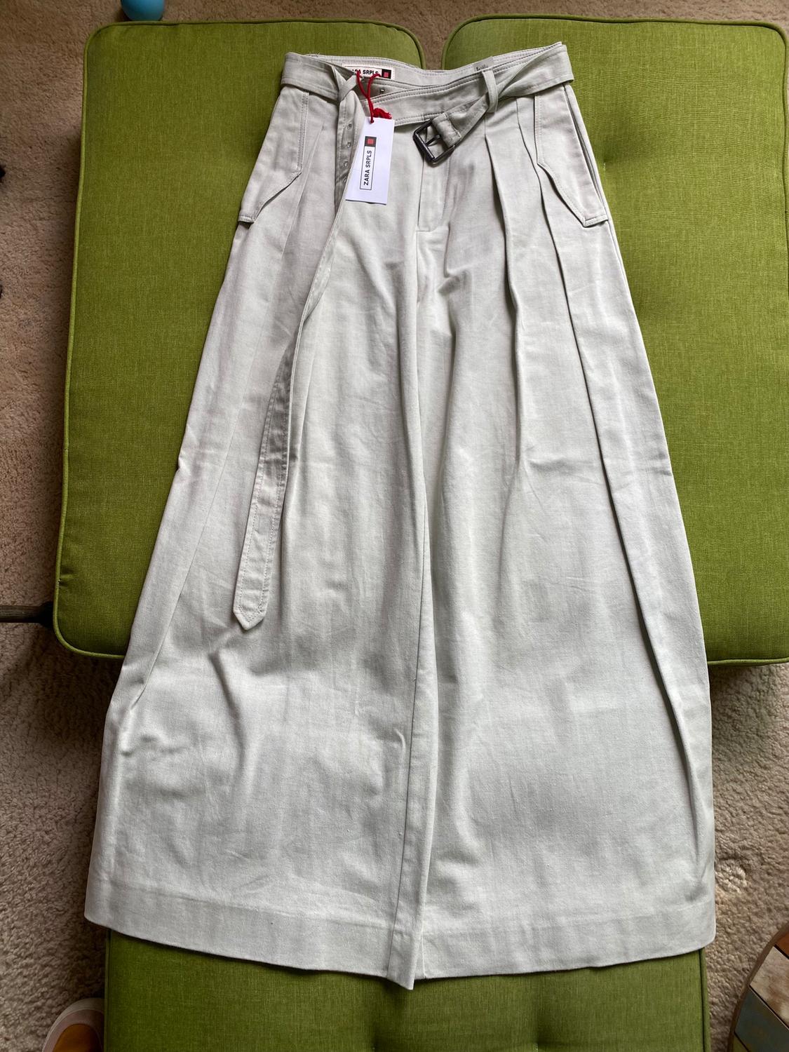 Beige pants with a belt and a white tag on a green sofa. Beige carpet background.