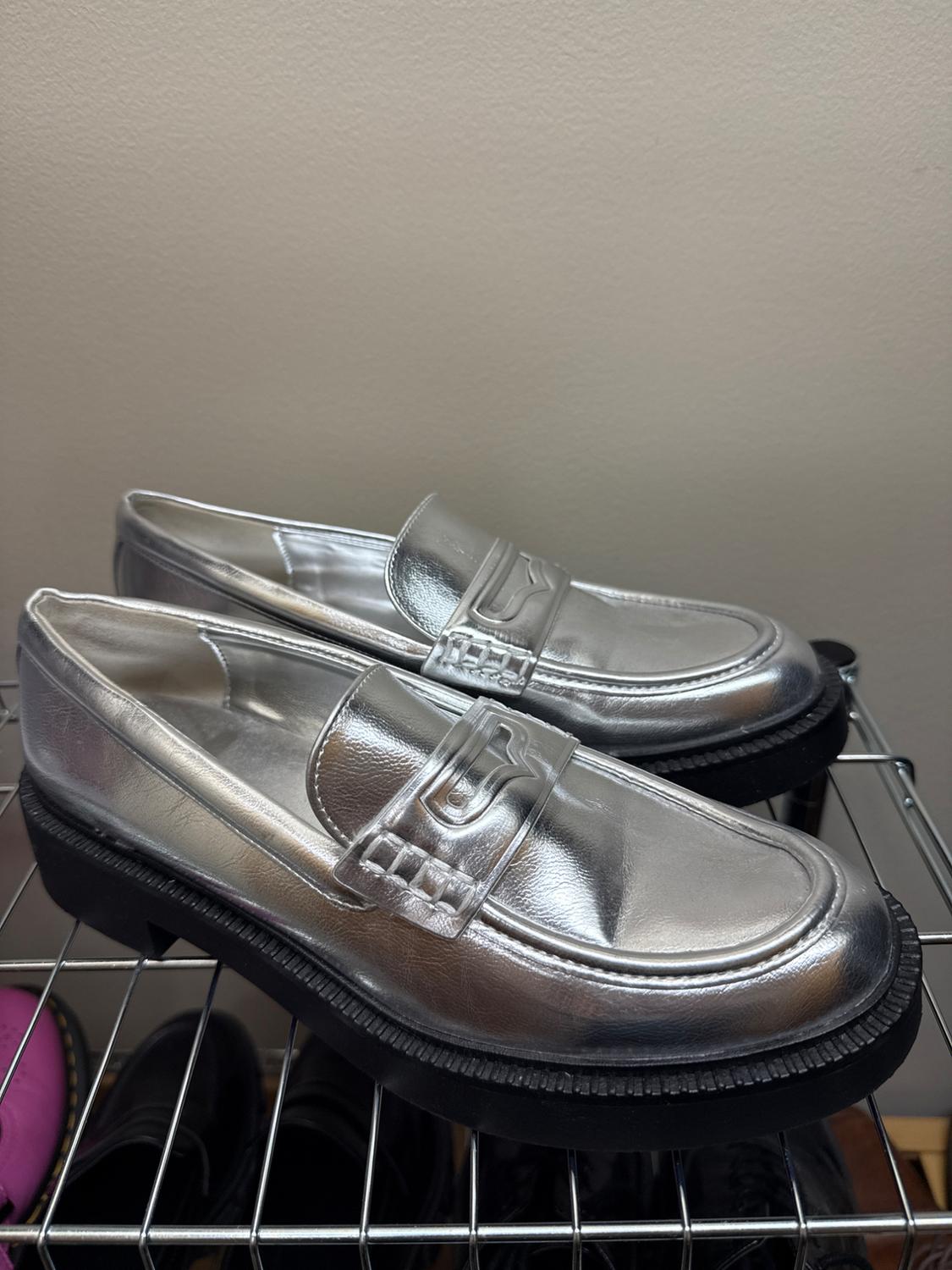 Silver patent leather shoes with a black sole on a metal grid. Beige background.