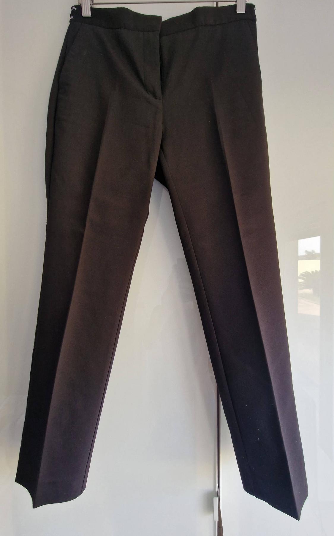 Dark formal trousers with a front crease, hanging from a hanger against a reflective white background.