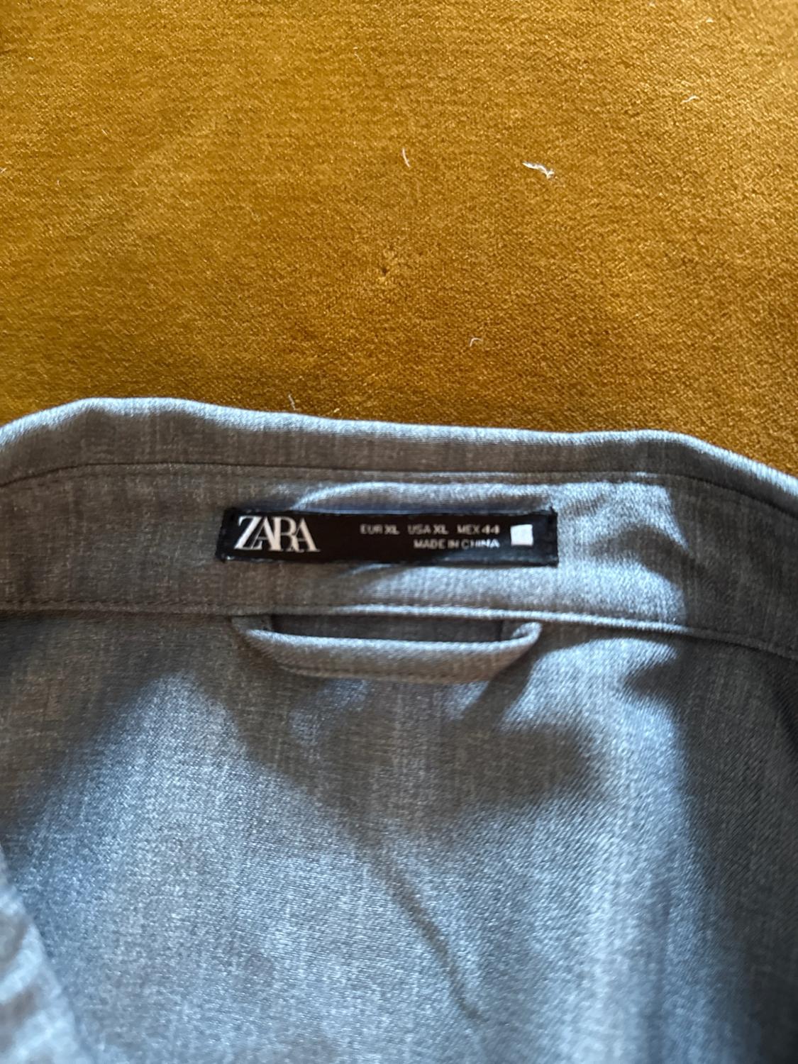 Close-up of a grey shirt collar against a mustard background. Black label with text: ZARA EUR XL USA XL MEX 44 MADE IN CHINA.