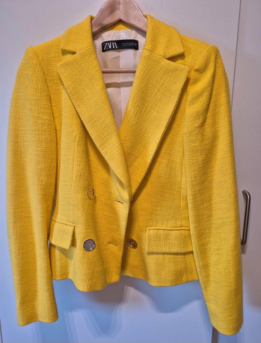Textured yellow jacket with buttons, hanging on a wooden hanger against a white door. Internal label with the text ZARA.