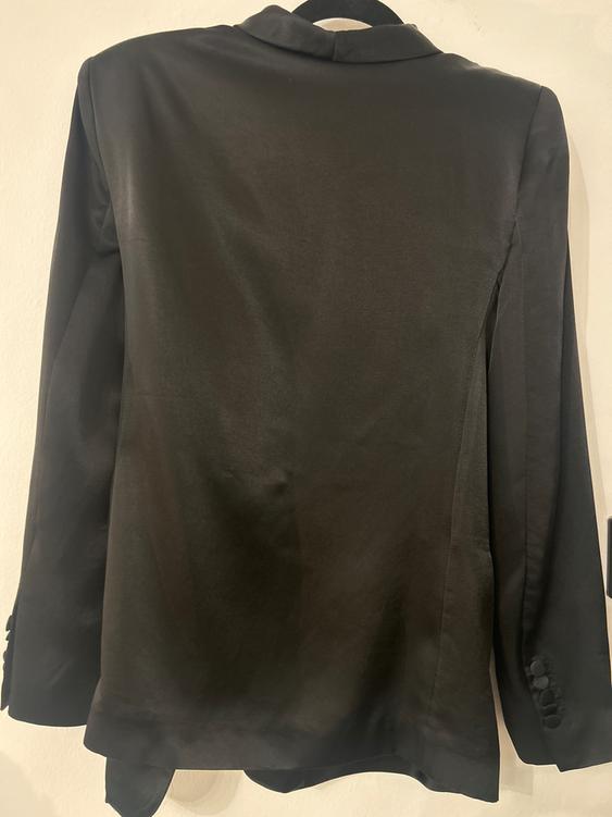 Black plain fabric jacket viewed from behind, hanging on a hanger against a white background. Long sleeves with buttons on the cuffs.