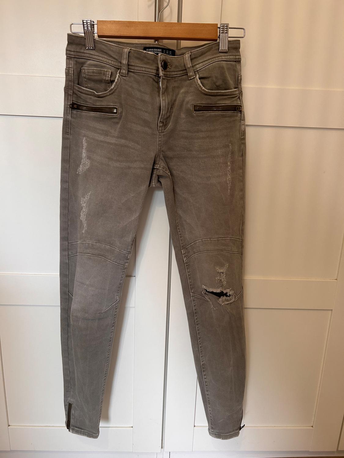 Worn grey trousers with zips on the front pockets and a rip at the knee, hanging on a wooden hanger.
