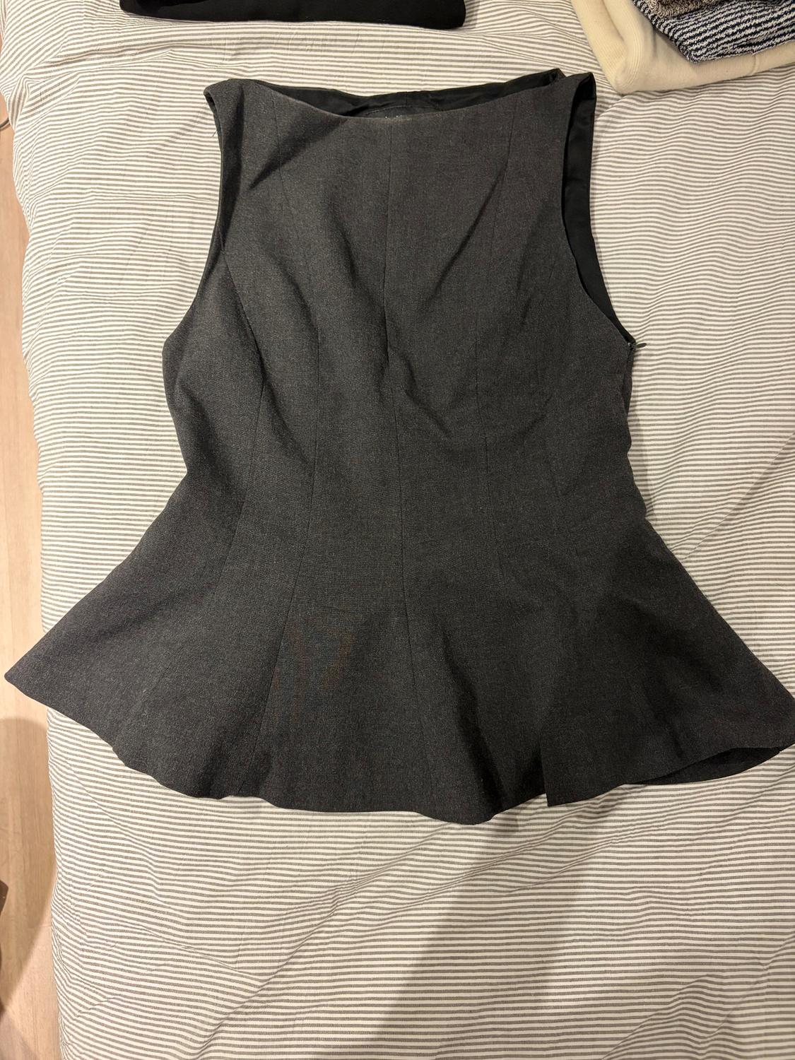 Dark grey sleeveless waistcoat on a bed with striped sheets. In the background, clothes folded in neutral colours.