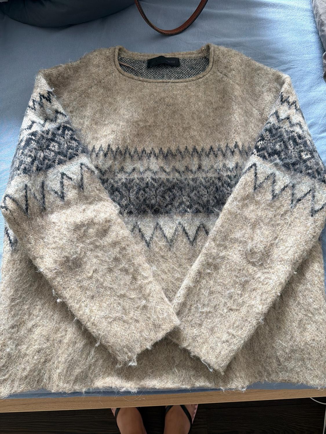 Beige knitted jumper with a grey geometric pattern on blue fabric. A handbag strap and some feet on a wooden floor are visible at the edges.