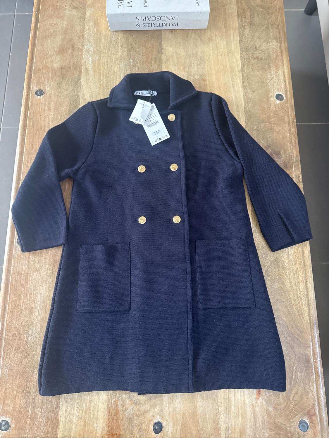 Navy blue coat with gold buttons and two front pockets on a wooden table. Tags hanging from the neck.