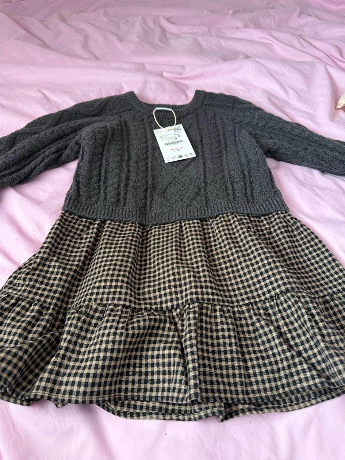 Dressed in formal wear on a pink sheet. Dark grey cable-knit top. Black and beige checked skirt with ruffles.