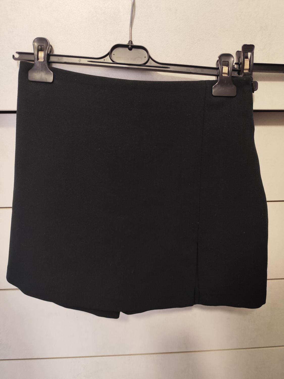 Black short skirt hanging on a black hanger with clips, against a light wooden background.