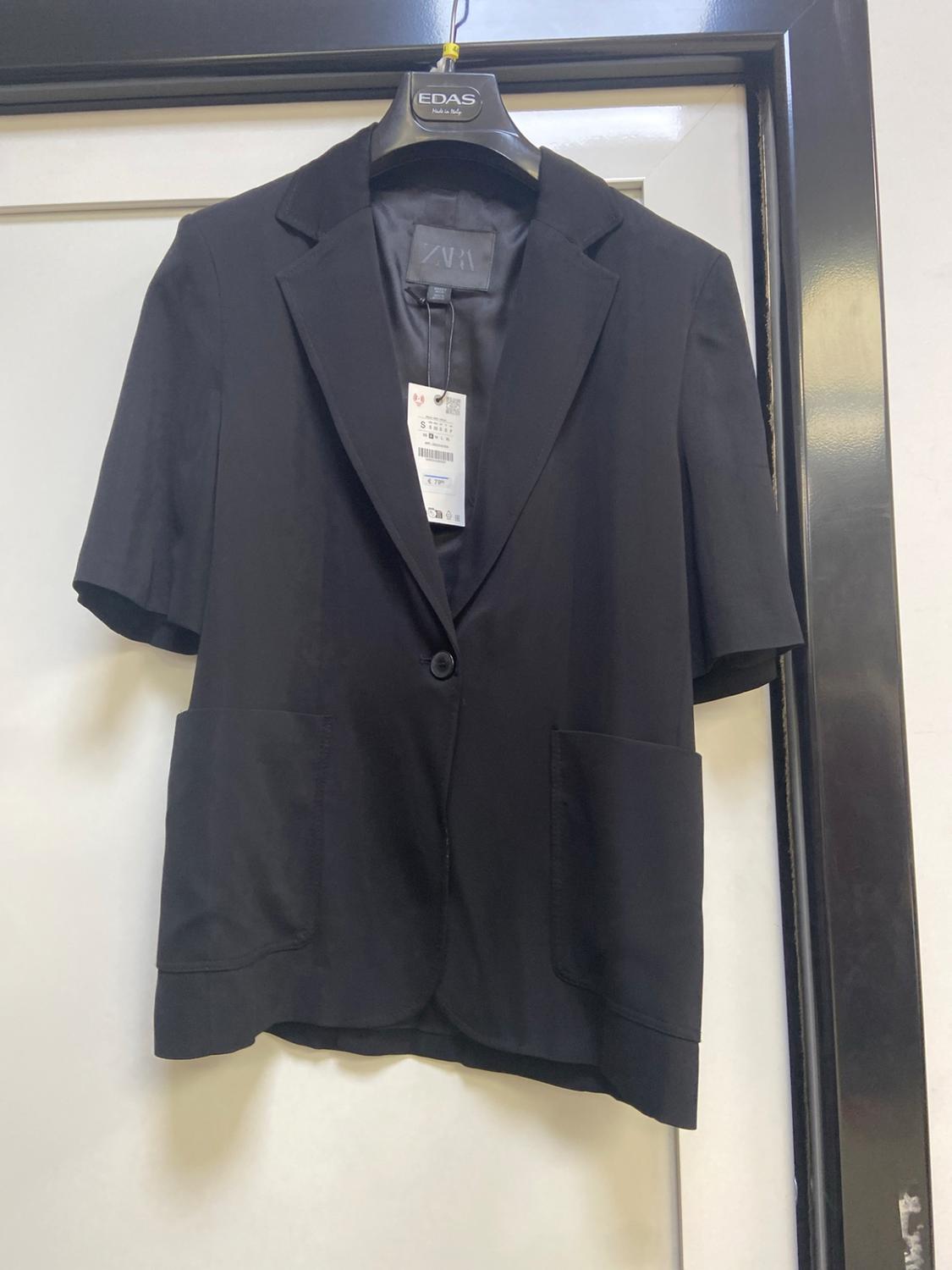 Black short-sleeved blazer with one button and two pockets, hanging on a hanger. Text: EDAS Made in Italy and ZARA.