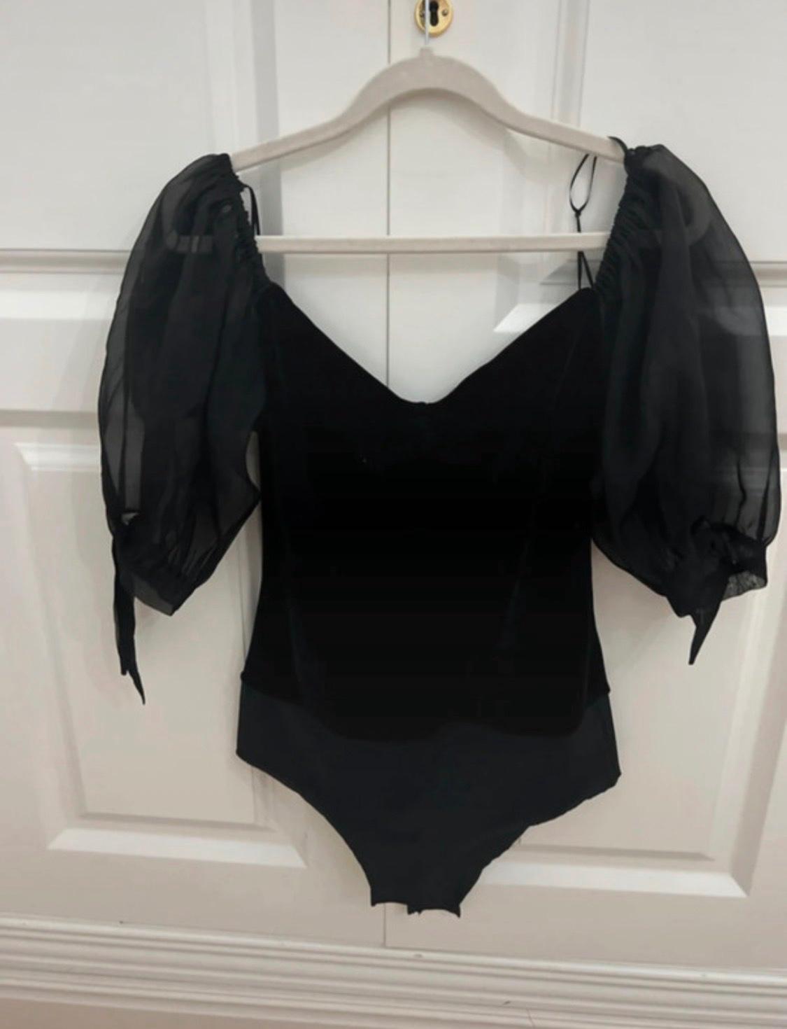 Black bodysuit with a V-neck and semi-sheer puff sleeves with ties, hanging on a hanger on a white panelled door.
