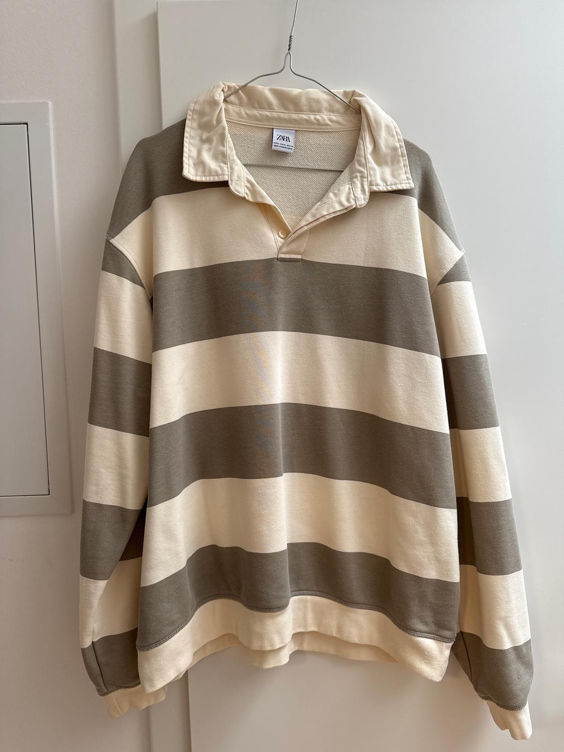 Wide-striped jumper in shades of beige and grey, hanging on a hanger. Visible label on the collar with the ZARA brand.
