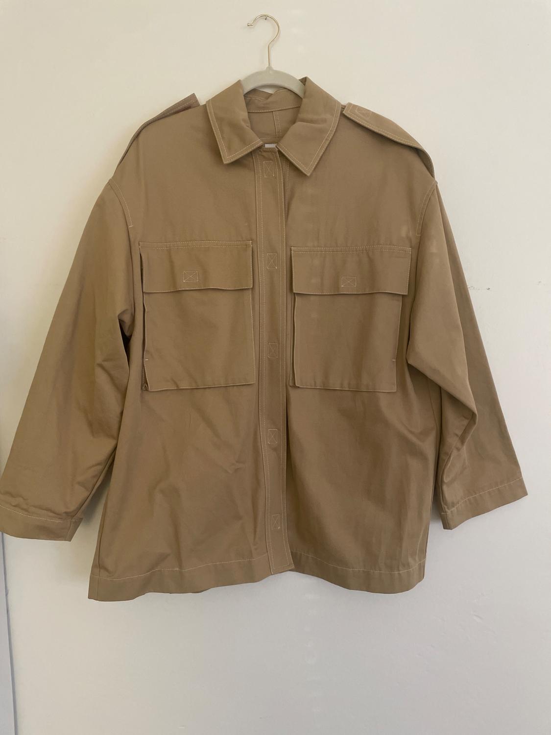 Long-sleeved beige jacket with large front pockets, hanging on a hanger against a white wall.