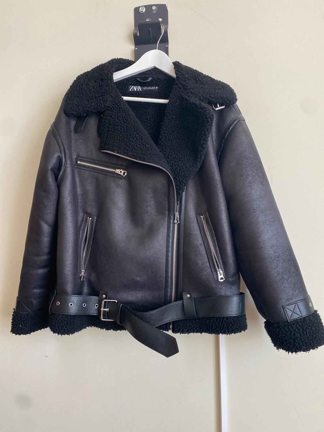 Black jacket with faux shearling lining, metal zips, and a belt, hanging on a white hanger. Visible label: ZARA.
