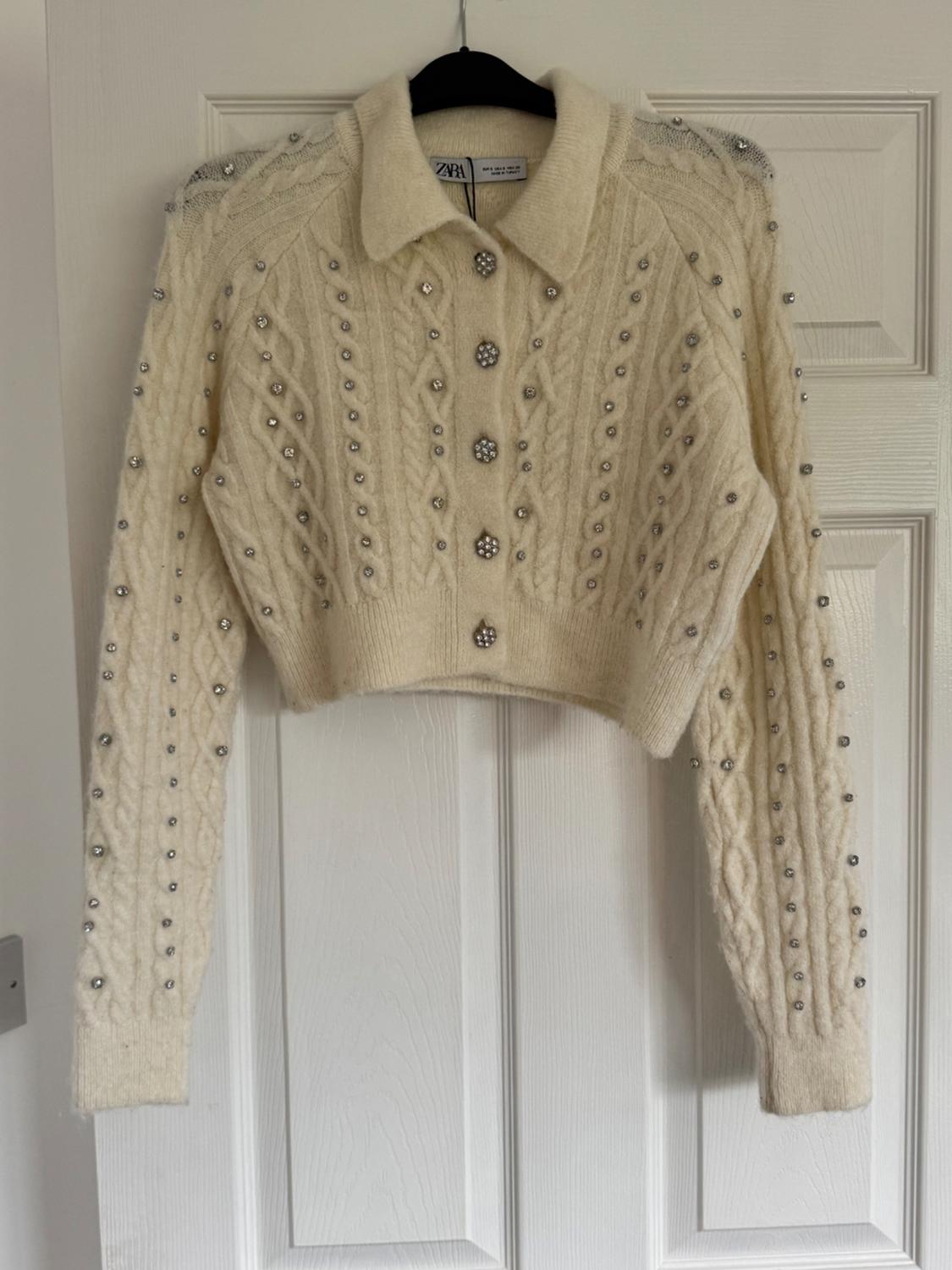 Short, cream-coloured knitted cardigan with a cable knit design, adorned with pearls and rhinestones, hanging on a hanger in front of a white door.